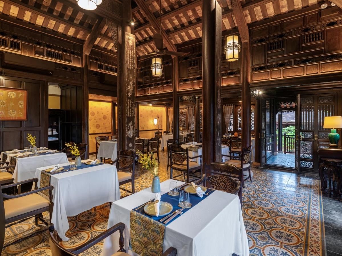 Dining room inside a garden house with dark wooden columns, patterned floor tiles, white tablecloths and curtained windows onto the garden