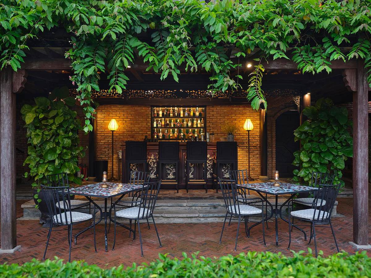 Garden bar under hanging green vines with a brick back wall, bottles on shelves, wall lamps and mosaic-topped tables