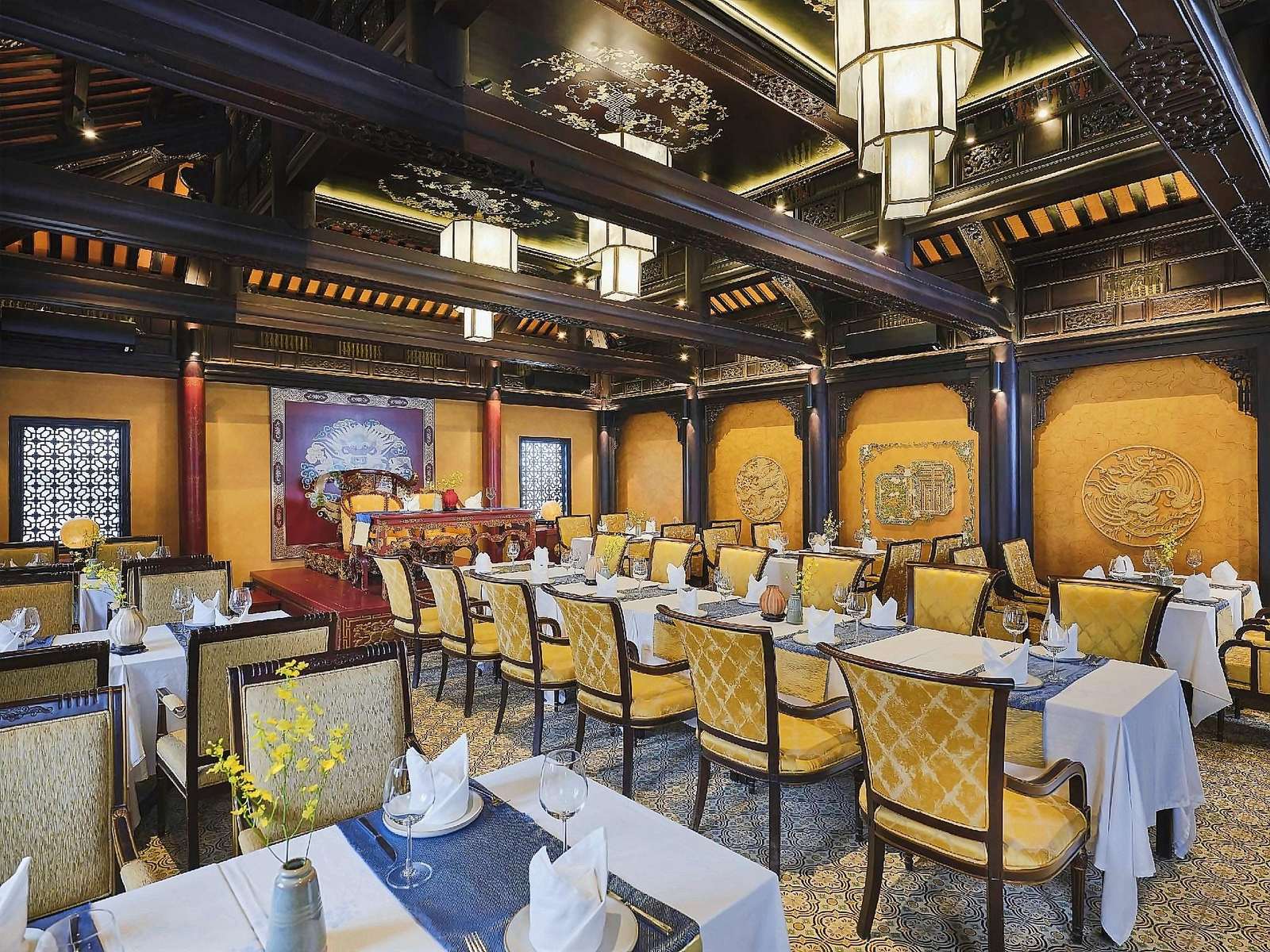 Dining hall with carved dark-wood beams, golden walls with round motifs, yellow upholstered chairs and tables laid with white cloths