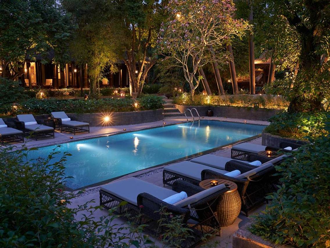 Rectangular pool at night lit blue, with sun loungers along its edge under trees and lanterns in the garden