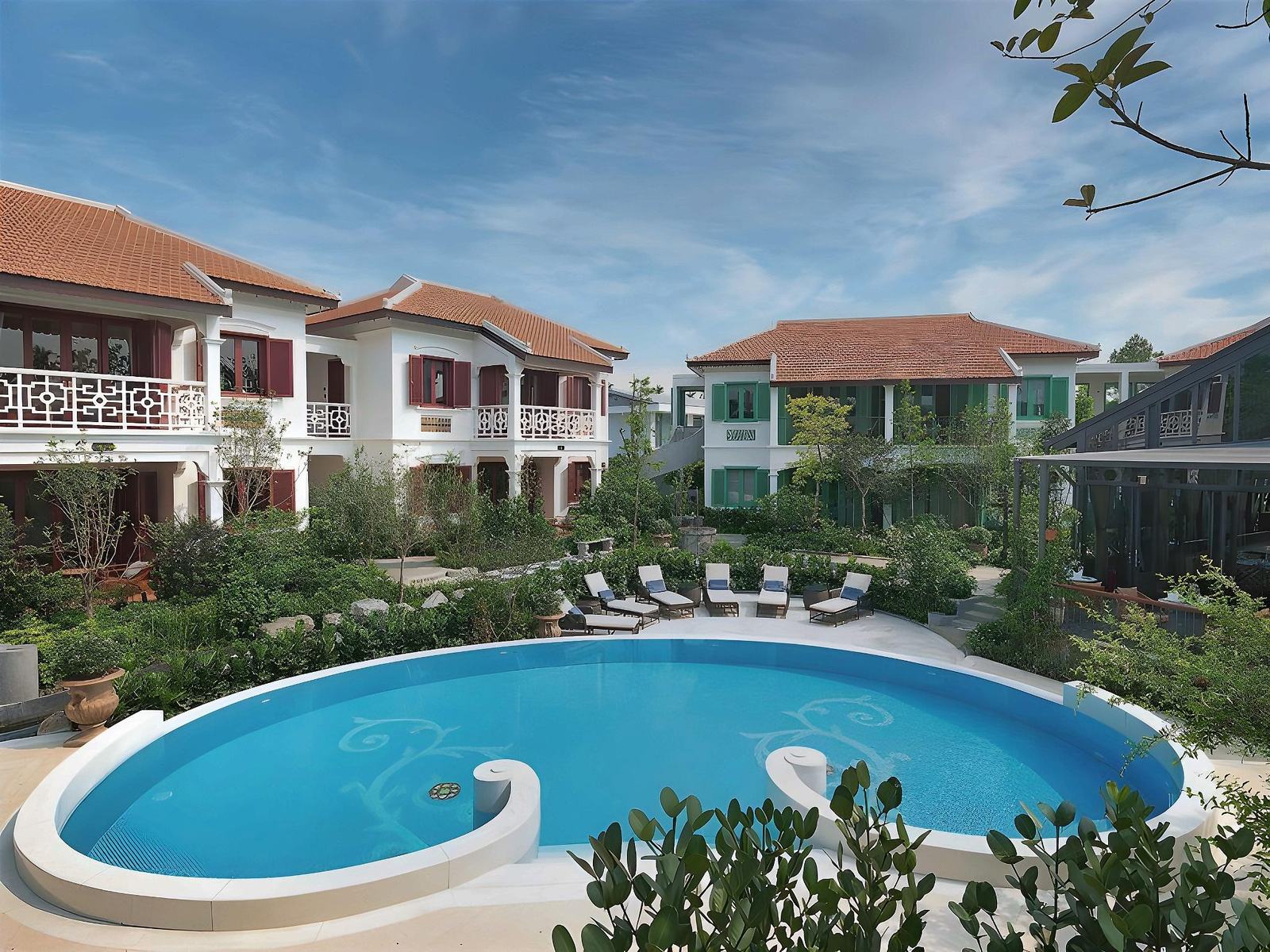 Curved blue pool with a round shallow section, sun loungers and umbrellas in front of two-storey cream villas with red tiled roofs