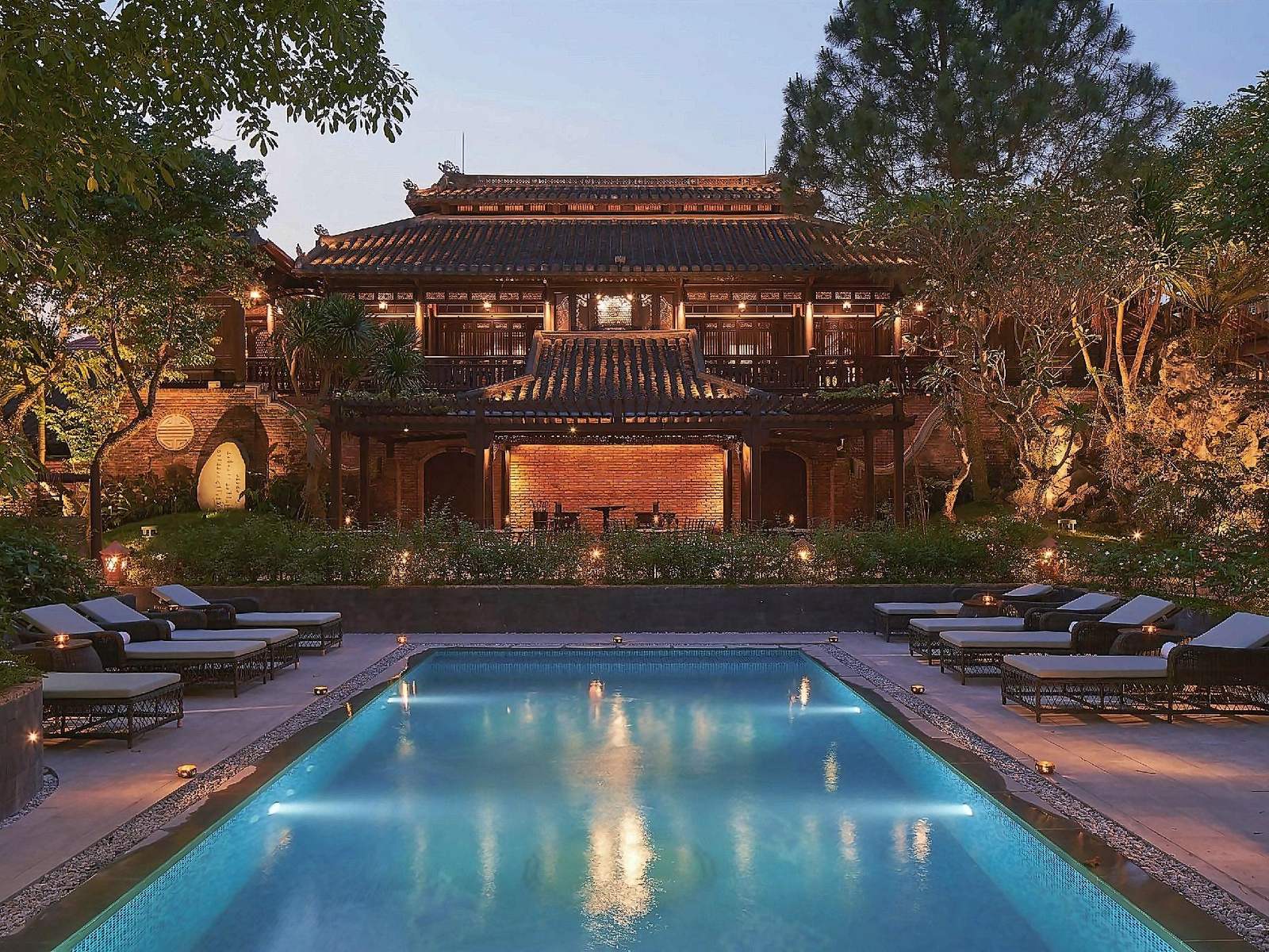 Aerial view at dusk of tiled villa roofs and dark wooden garden houses around a lit pool, with a residential neighbourhood and trees beyond