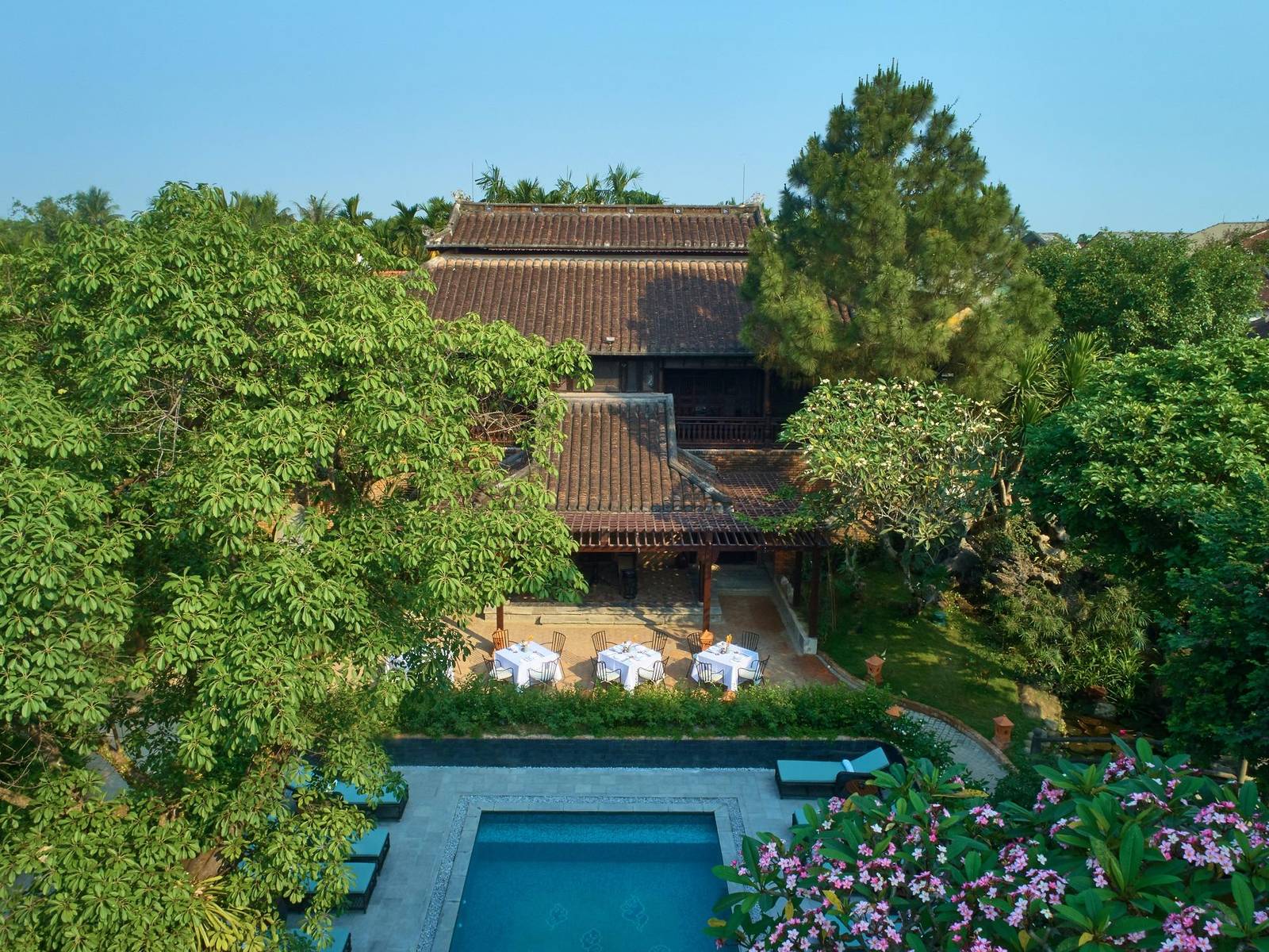 Aerial view of a dark-wood garden house with tiled roofs among trees, a pool with loungers in front and pink flowers in the garden