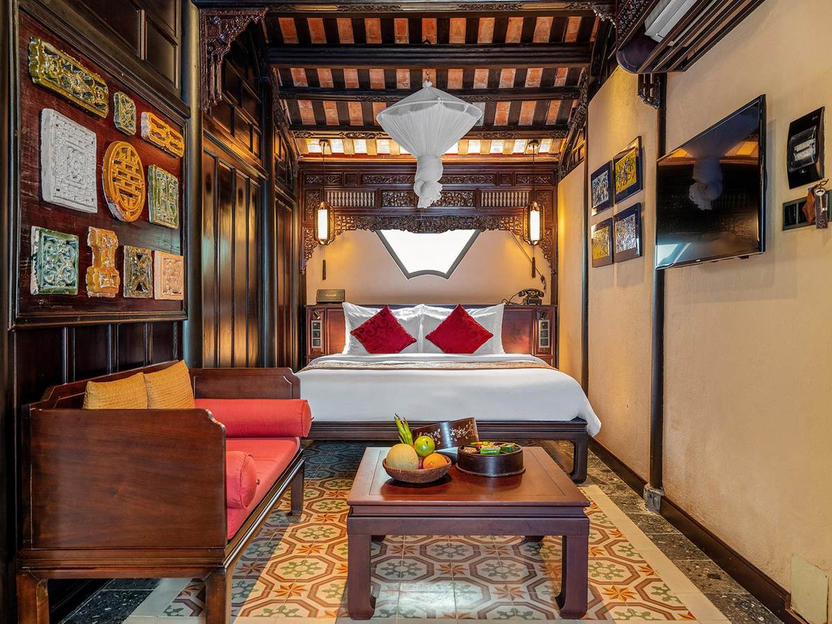 Garden-house bedroom with a dark carved wooden frame, a daybed with red cushions, patterned floor tiles and a white canopy over the bed