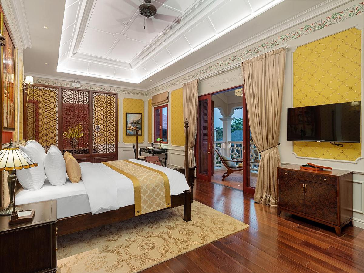 Large villa room with yellow walls, dark wooden floor, a bed with a gold runner and tall windows with long curtains