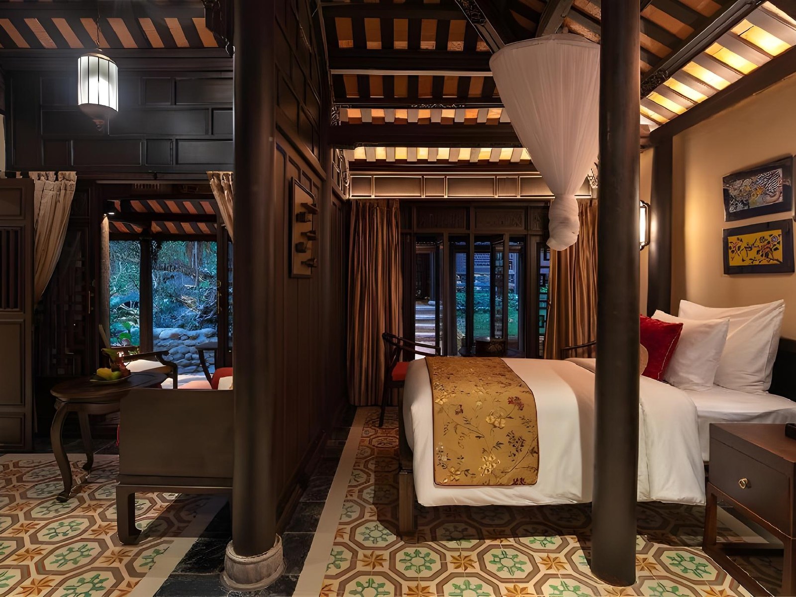 Garden-house bedroom seen through dark wooden pillars, with a bed under a white canopy, patterned floor tiles and a green garden beyond the glass doors